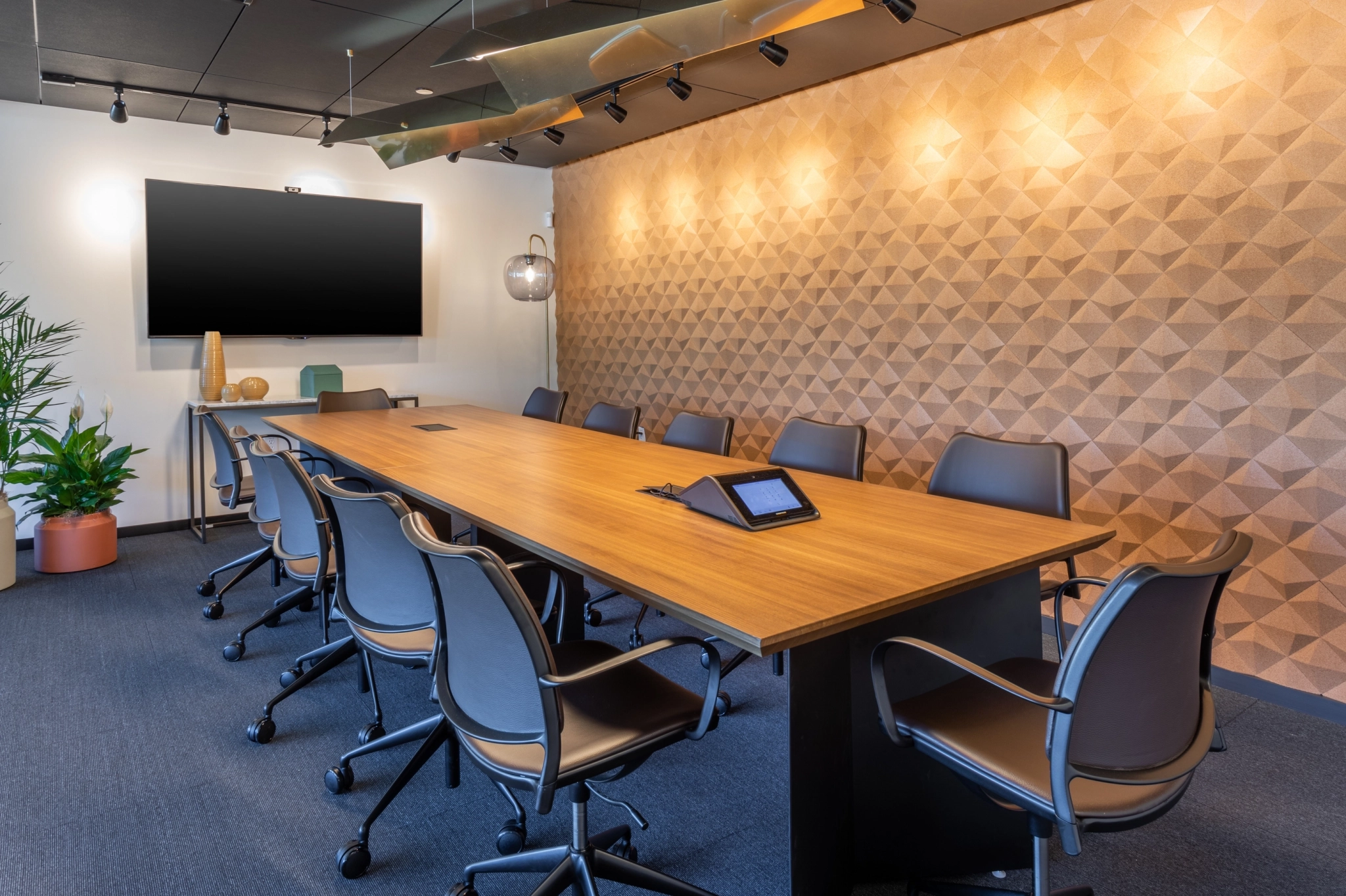 Modern meeting room in Phoenix featuring a long wooden table, ten chairs, a wall-mounted screen, potted plants, and geometric-patterned wall panels—ideal for coworking sessions and collaborative gatherings.