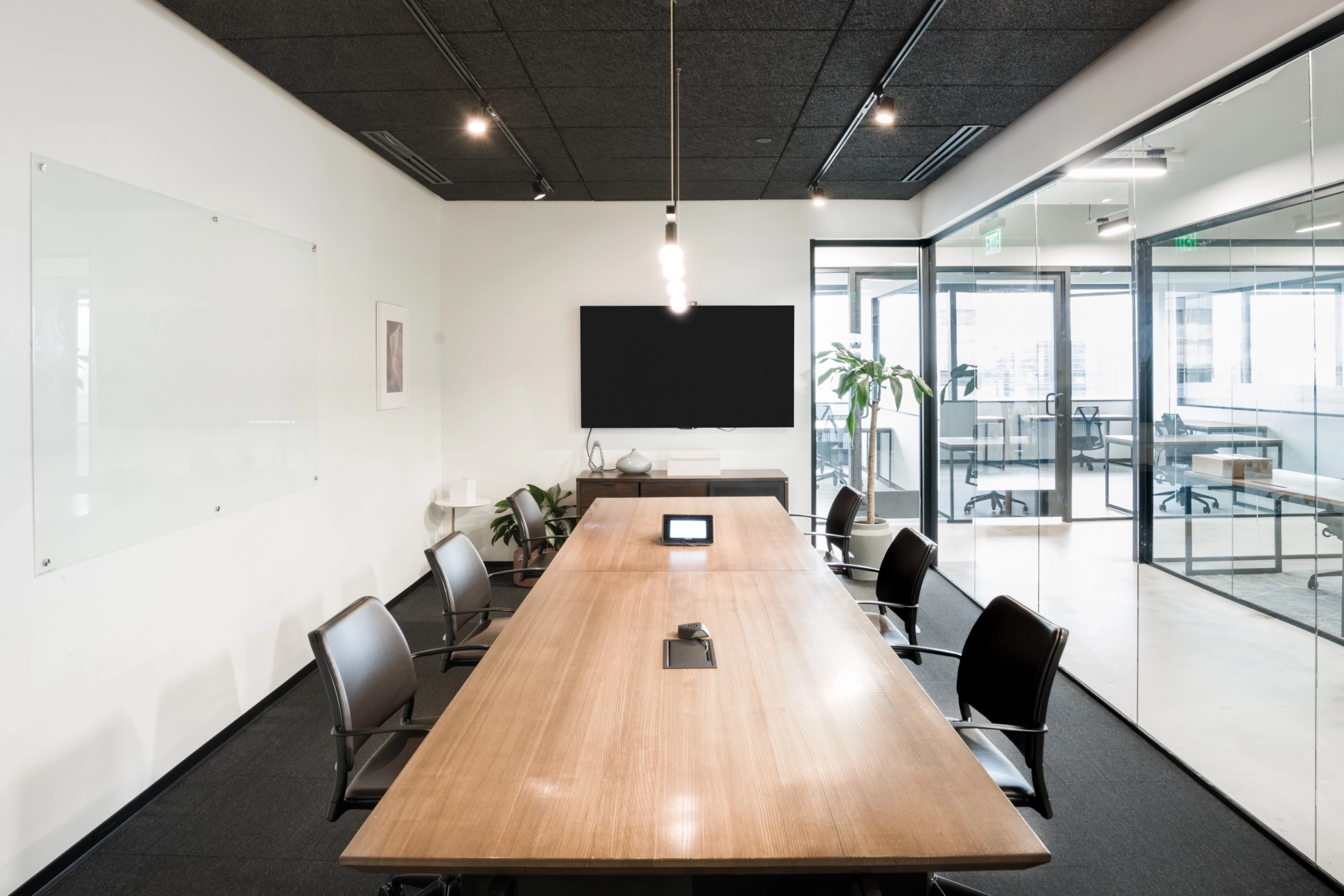 Salle de réunion moderne à Phoenix comprenant une longue table en bois, six chaises noires, un grand écran, un tableau blanc et des murs en verre avec vue sur l'espace de coworking.