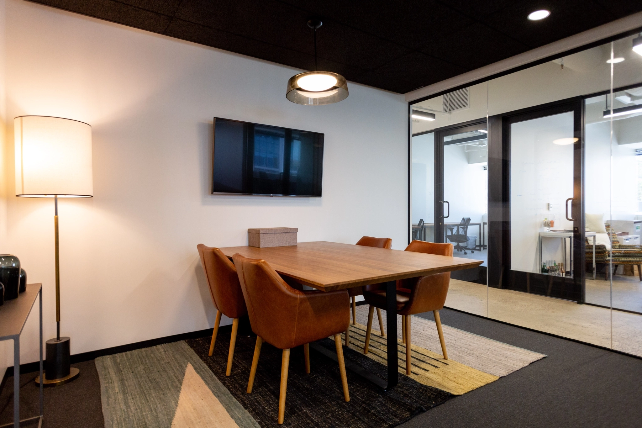 A small conference room in a Phoenix coworking space, featuring a wooden table, four brown chairs, a wall-mounted TV, floor lamp, and glass walls overlooking the adjacent office workspace.