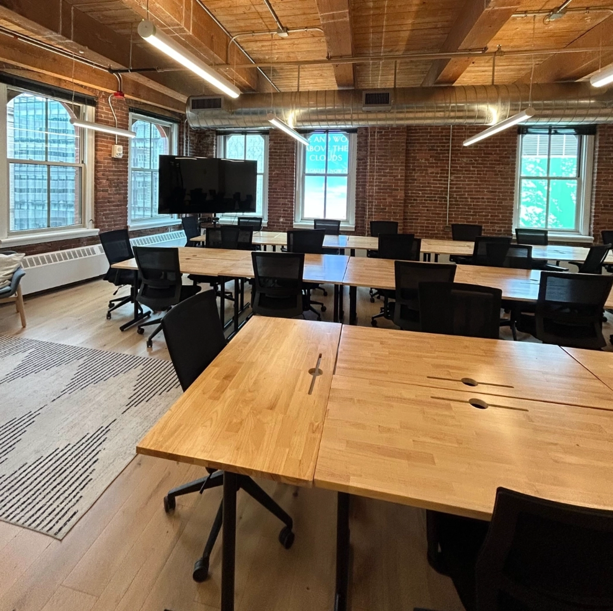 A modern coworking space in Boston featuring several wooden desks, black chairs, large windows, exposed brick walls, a TV screen, and visible ceiling pipes.
