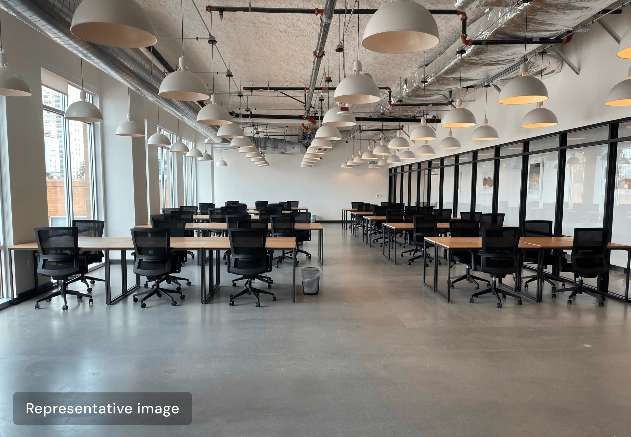 A modern open-plan office workspace with rows of empty desks and black chairs, large windows on one side, and white pendant lights hanging from the ceiling.