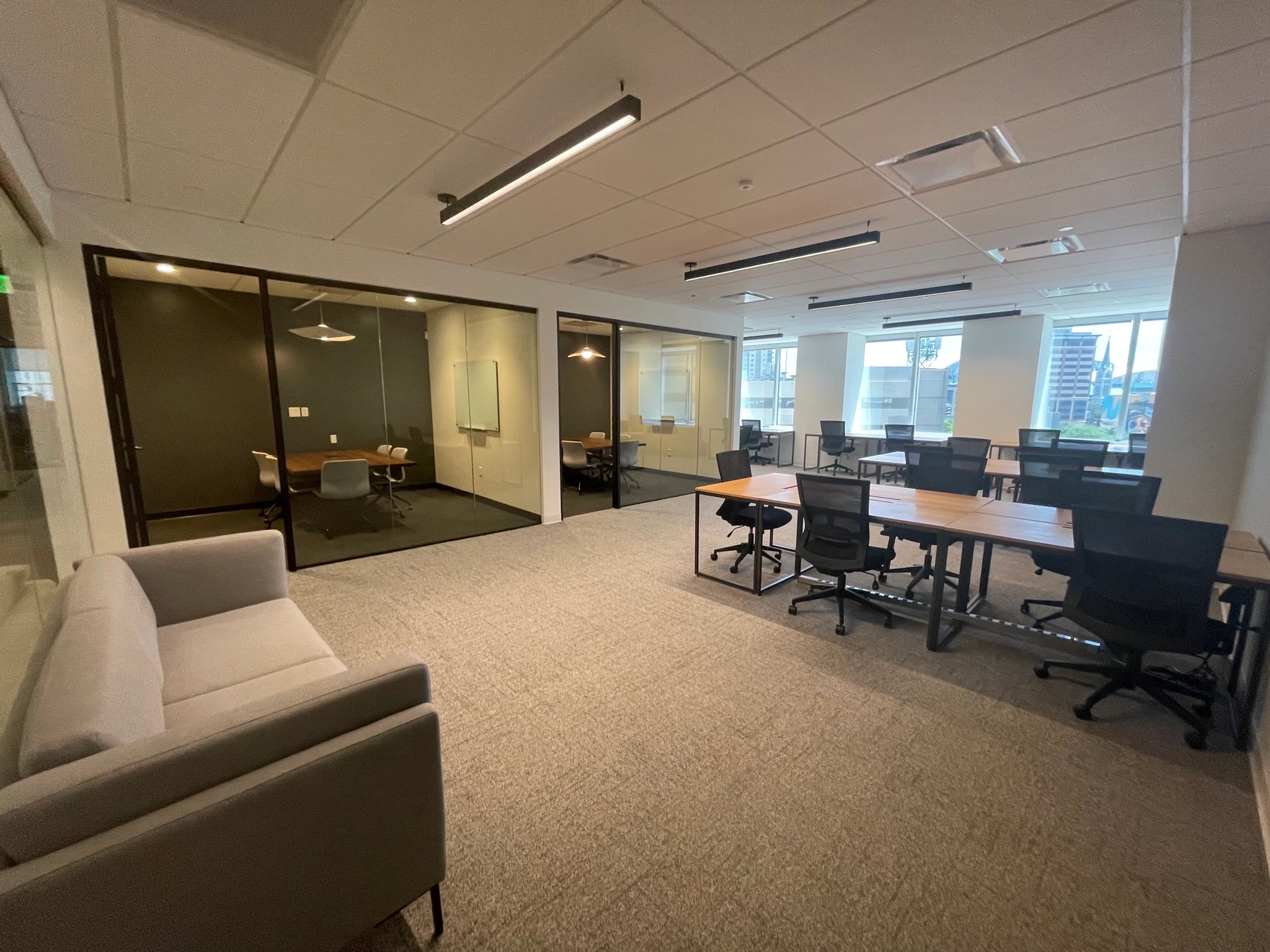 Modern Houston office space featuring open desks, black office chairs, glass-walled meeting rooms, a gray sofa, and large windows providing natural light.