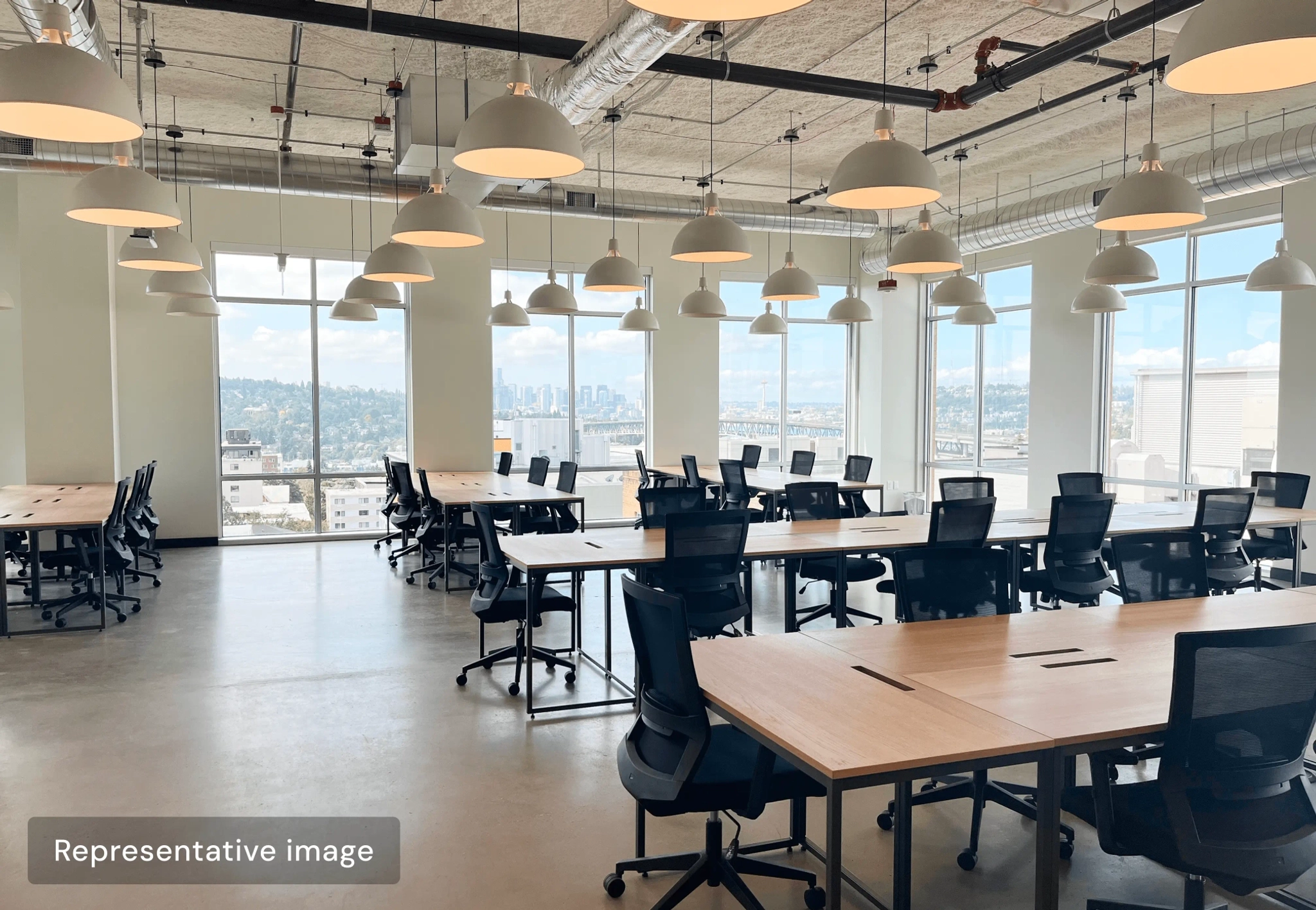 Modern open-plan office with several empty desks and chairs, large windows offering a New York city view, and ceiling-mounted pendant lights.