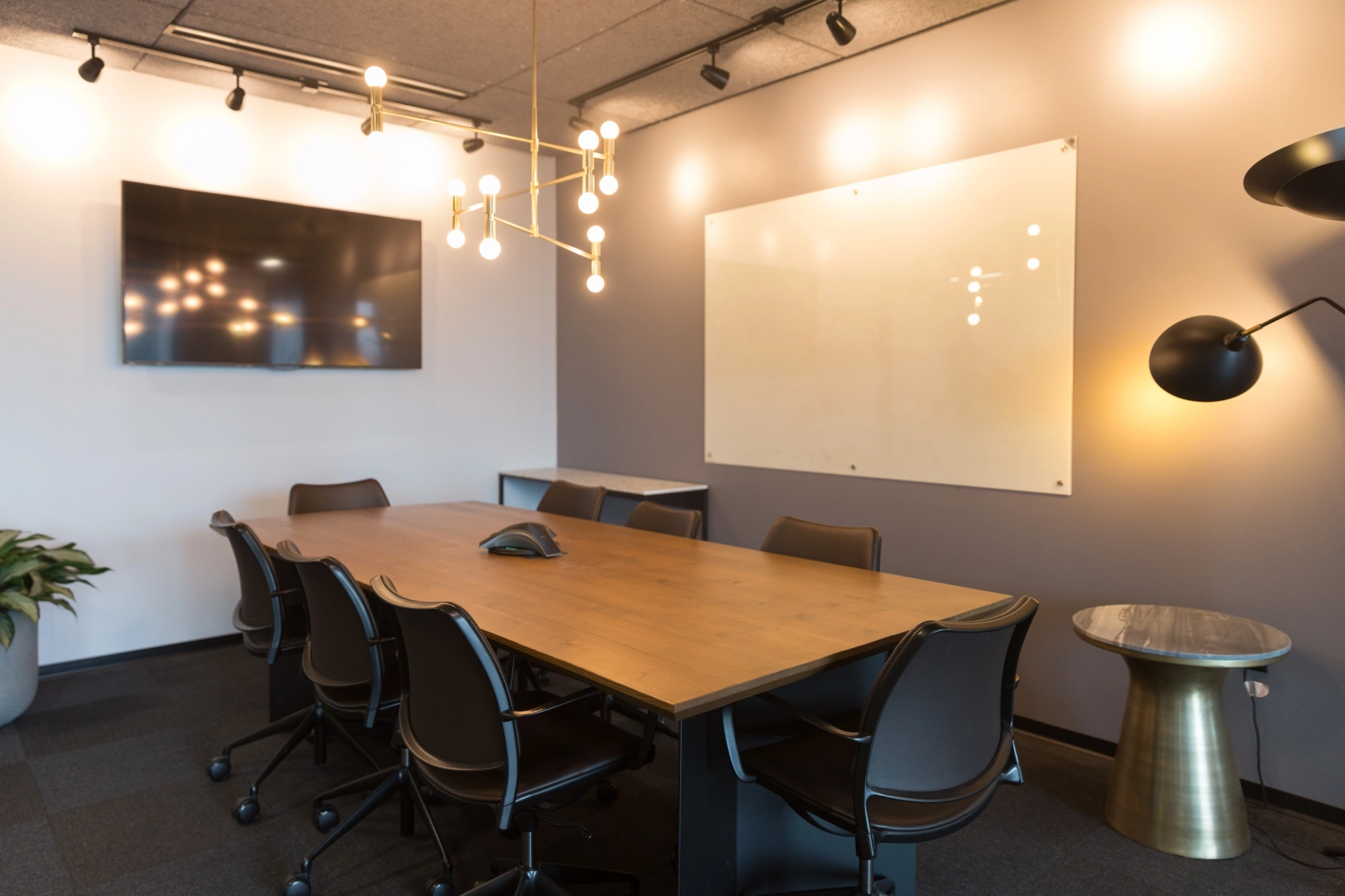 Modern meeting room with a wooden table, eight chairs, wall-mounted TV, whiteboard, stylish light fixtures, and a small side table—perfect for any collaborative workspace.