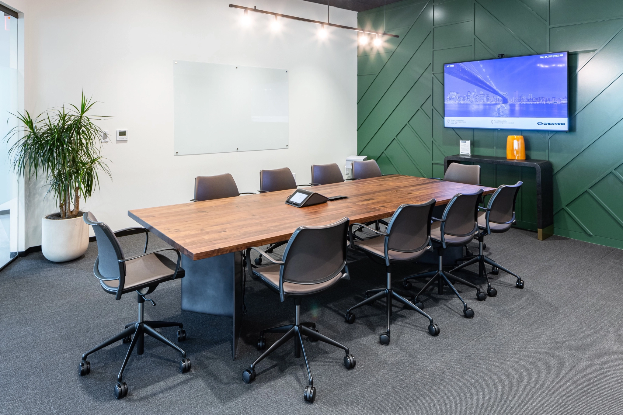 Modern meeting room with a large wooden table, eight chairs, a wall-mounted screen, whiteboard, potted plant, and a geometric green accent wall—perfect for any collaborative workspace.