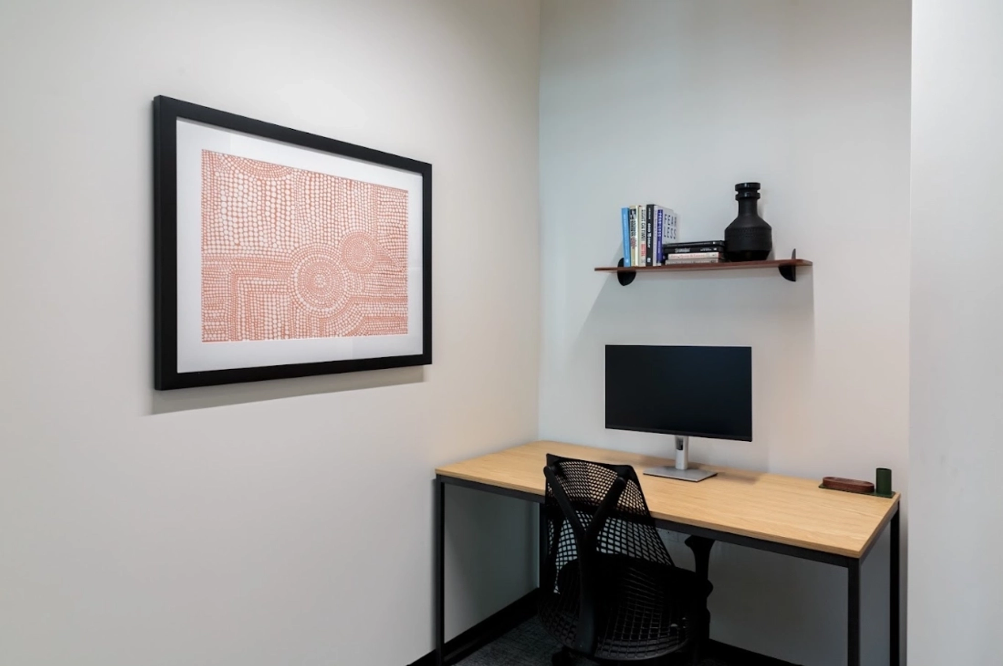 A small office workspace with a desk, computer monitor, office chair, a floating shelf with books and decor, and framed abstract art on a white wall—perfect for focused work or as an inviting meeting room corner.