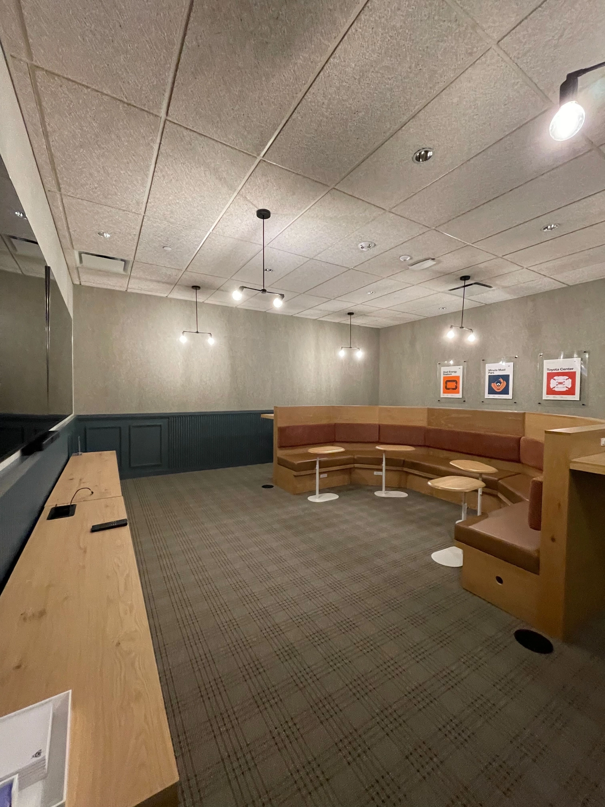 A modern coworking lounge area with brown cushioned seating, small round tables, pendant lights, and framed posters on the wall. The empty space features neutral-colored decor—ideal for a relaxed Houston workspace.