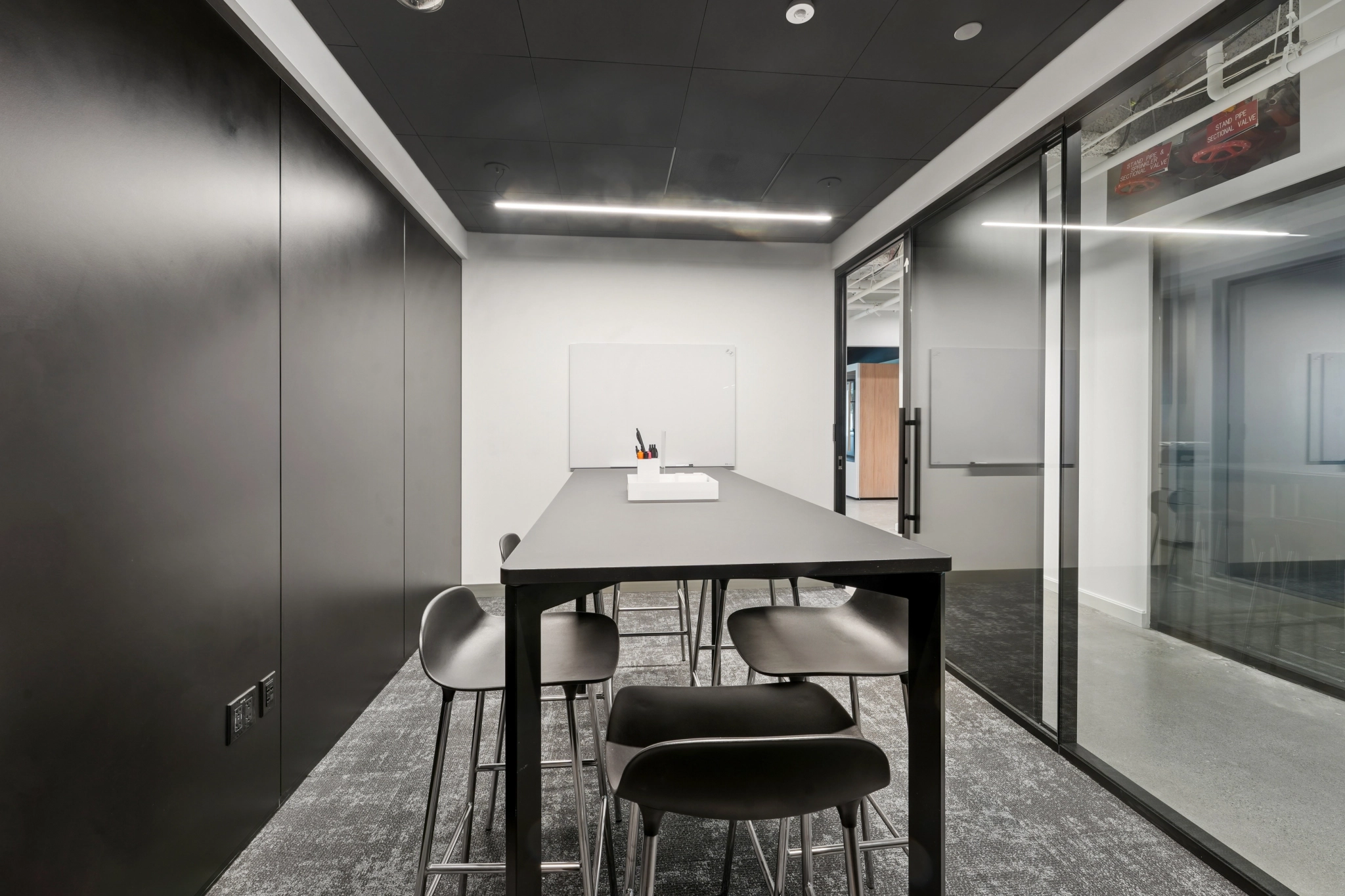Modern coworking meeting room with a long black table, six black chairs, glass walls, and a whiteboard on the far wall. A tray with pens and paper is on the table.