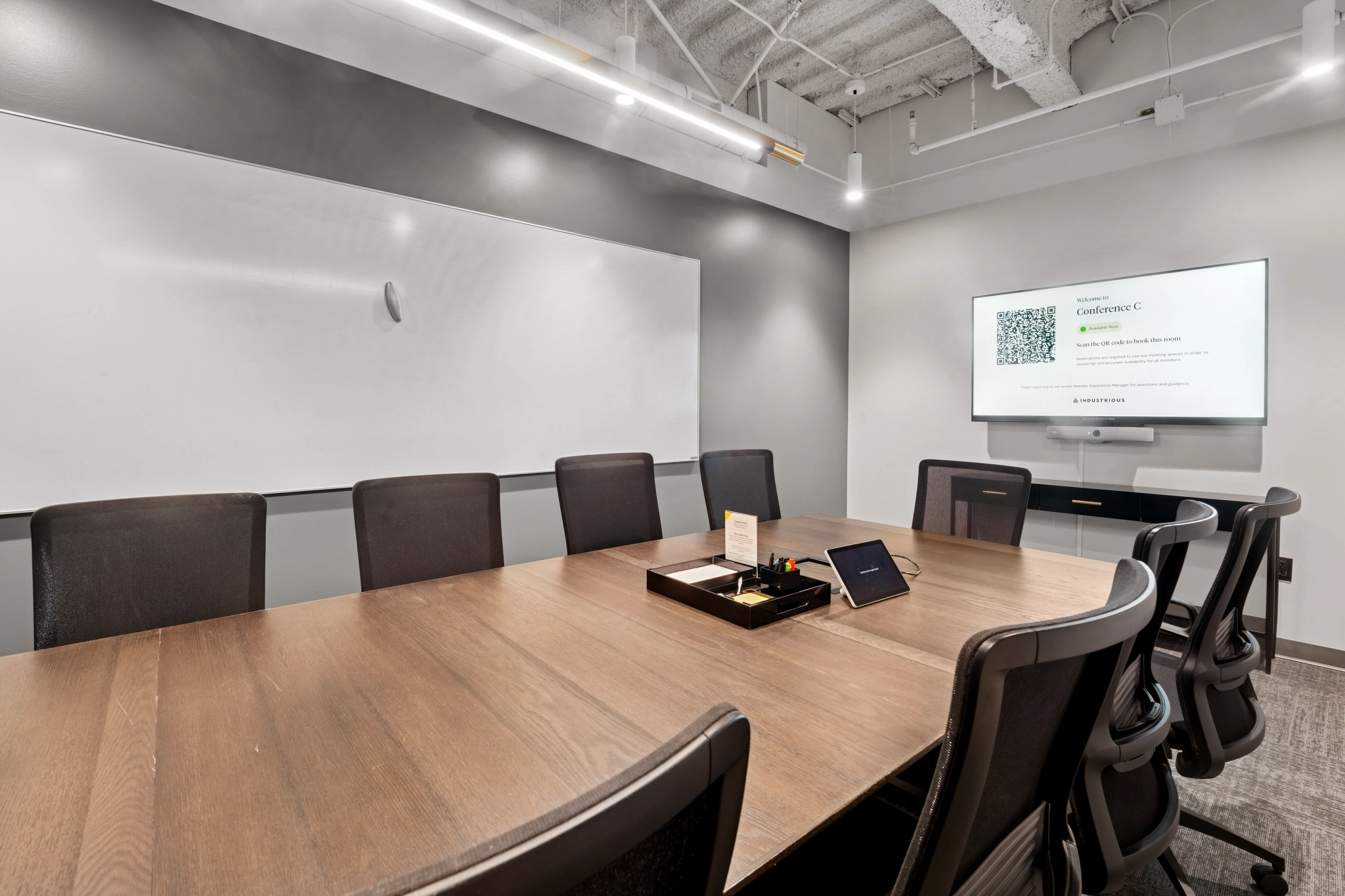 Modern Seattle meeting room featuring a long wooden table, black office chairs, a large whiteboard, and a wall-mounted screen displaying a QR code and conference information.