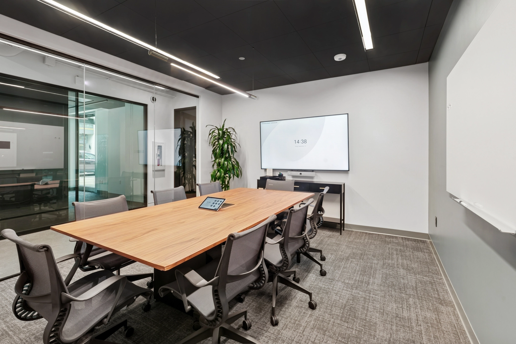 Modern coworking conference room with a wooden table, eight office chairs, a large screen on the wall, a whiteboard, a plant, and a glass wall to the left—perfect for your next meeting.