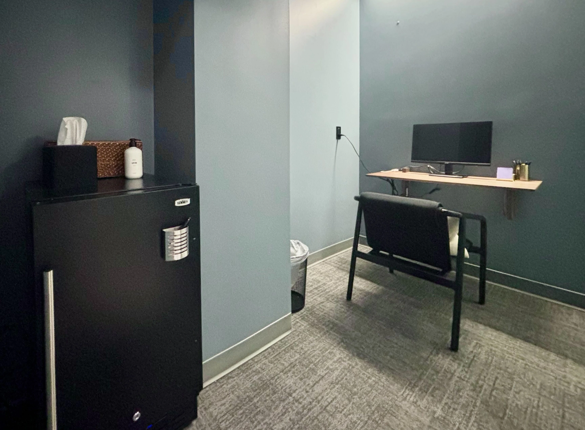 Un petit espace de travail à Seattle avec un bureau, un écran, une chaise et des articles de papeterie. Un mini-frigo, des mouchoirs en papier et une bouteille de désinfectant sont posés contre un mur gris. Le sol recouvert de moquette et les murs de couleur neutre créent un environnement confortable.