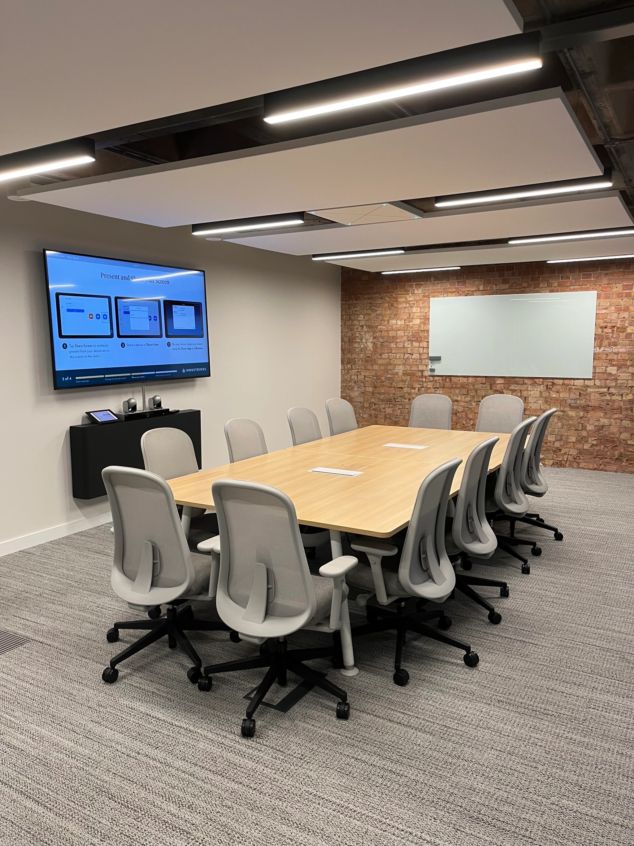 A modern coworking conference room with a large wooden table, ten gray chairs, a wall-mounted screen displaying presentations, a whiteboard, and a brick accent wall.