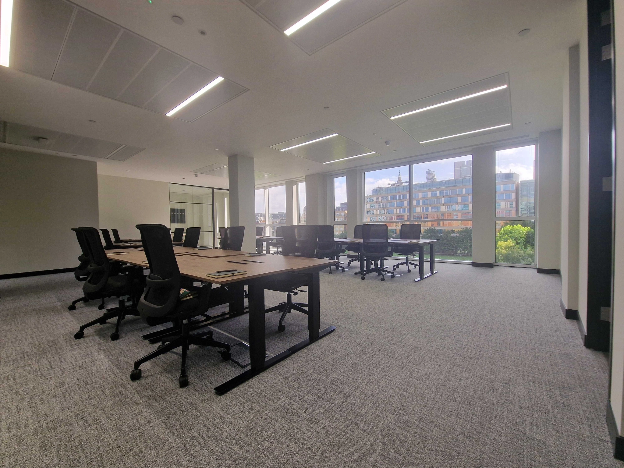 A modern office space with several desks and chairs arranged in rows near large windows overlooking buildings and greenery in London.