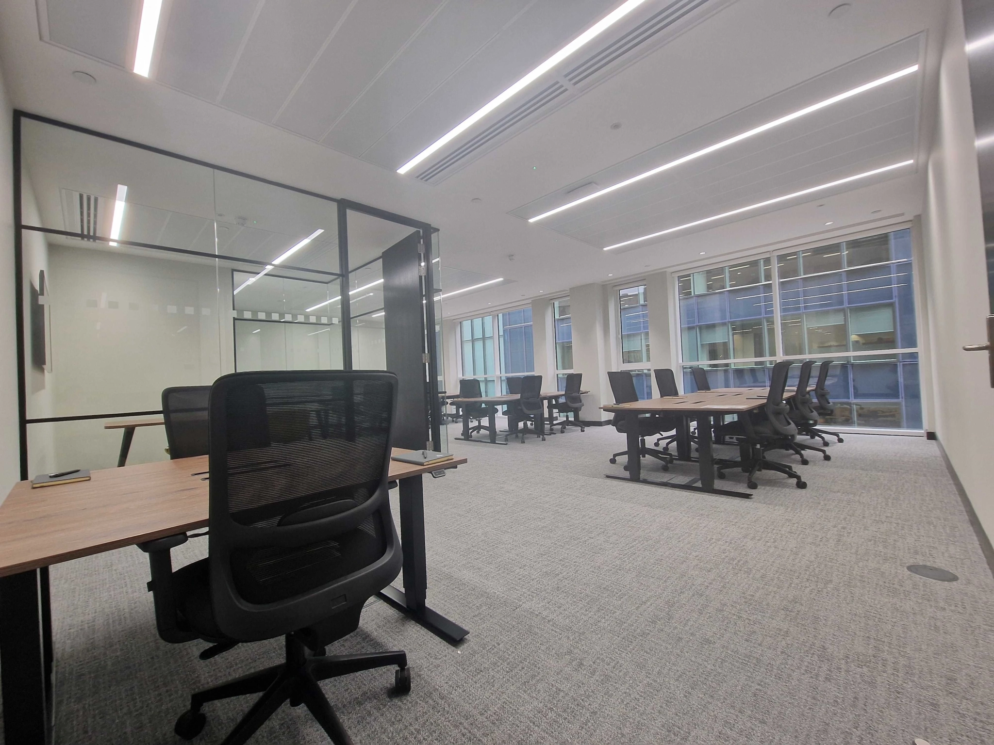 Modern coworking office space in London, featuring several desks and black chairs, large windows, a glass-walled meeting room, and bright overhead lighting.