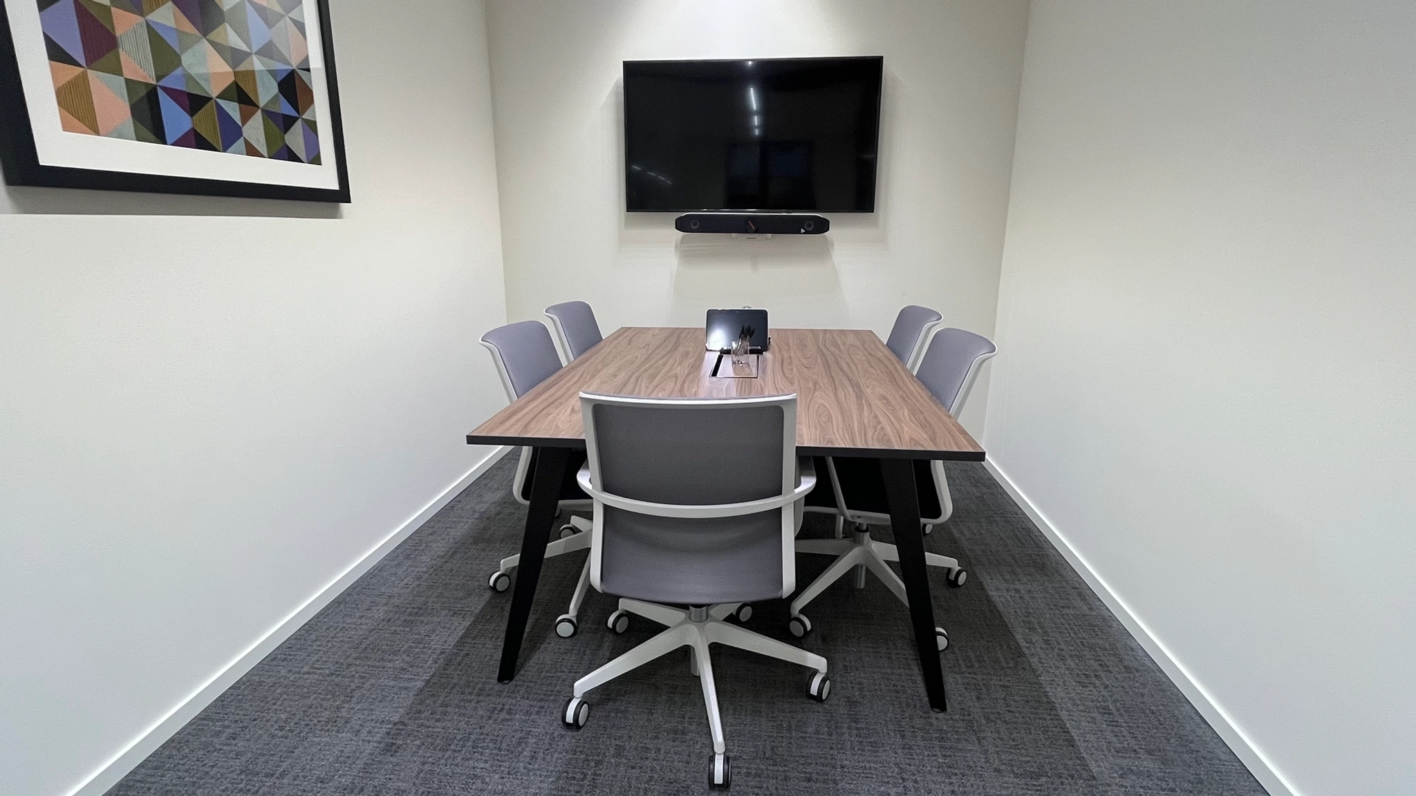 A small meeting room with a rectangular table, five chairs, a wall-mounted TV, and a framed abstract artwork on a white wall—perfect for focused coworking sessions or office discussions.