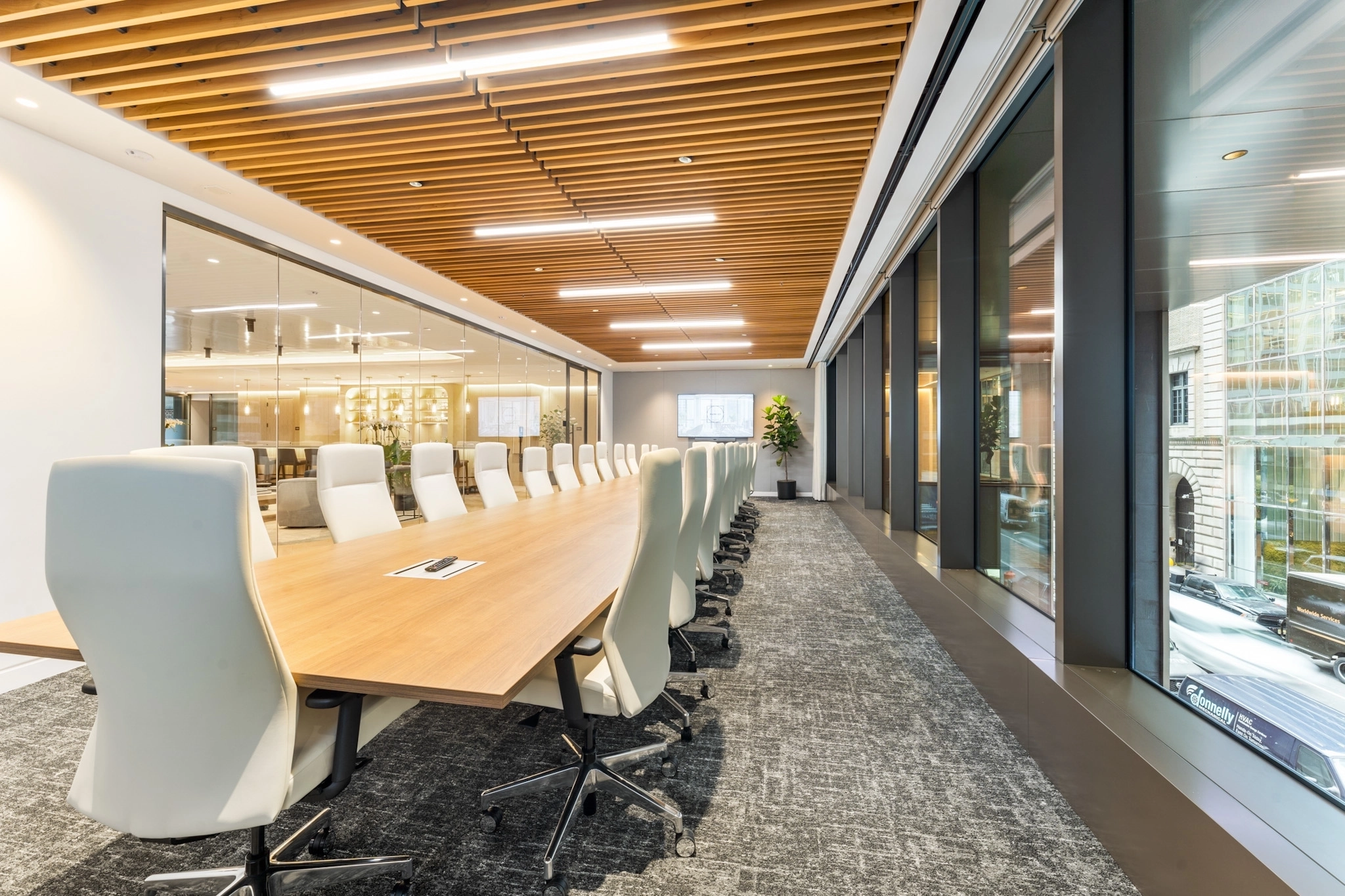 Modern meeting room with a long wooden table, white chairs, carpeted floor, wood slat ceiling, and large windows overlooking a vibrant New York street.