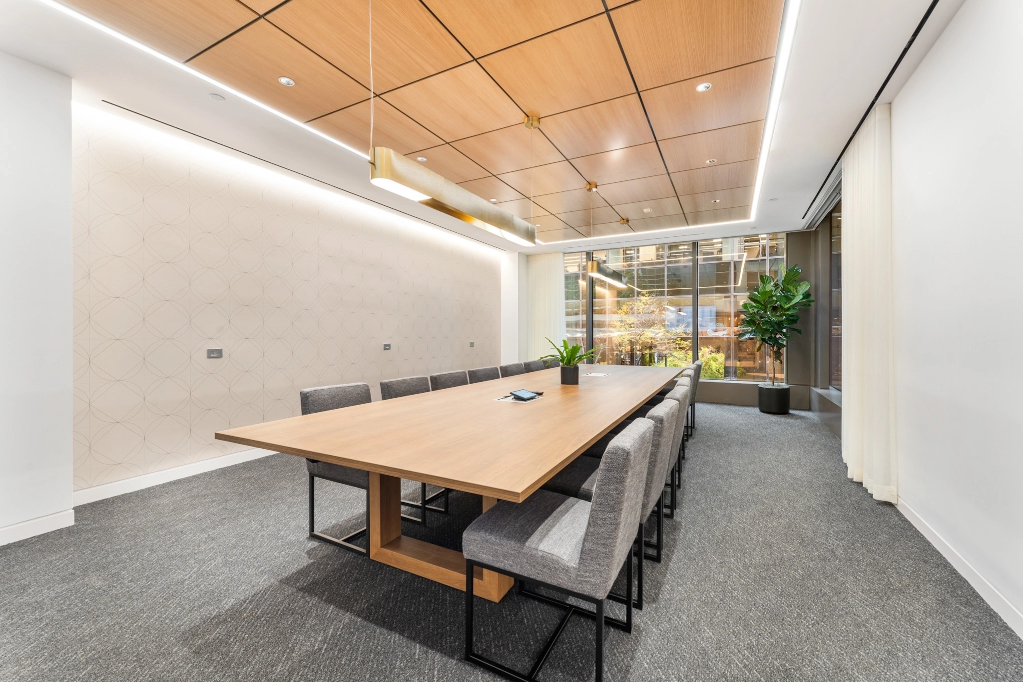 A modern office meeting room with a long wooden table, gray chairs, overhead lighting, carpeted floor, large windows, and indoor plants.