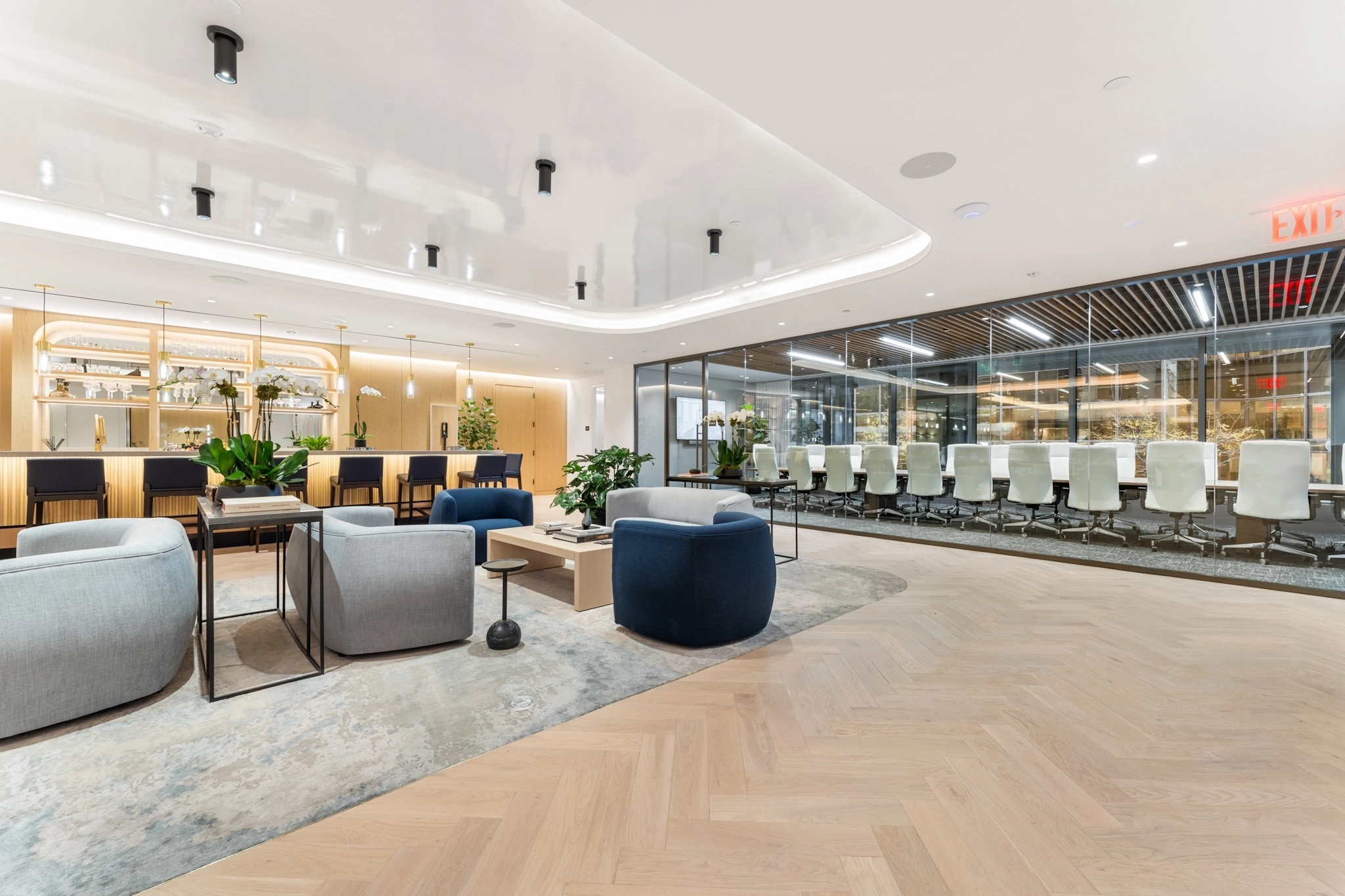 Modern coworking office space with lounge seating, a large meeting room featuring white chairs, light wood flooring, and glass walls that separate the conference area from the lounge.