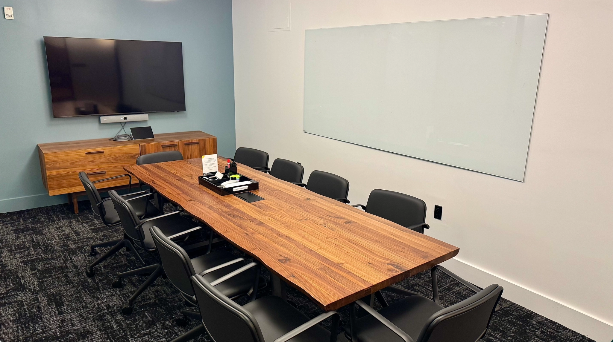 A modern coworking conference room in New York featuring a wooden table, ten chairs, a large wall-mounted screen, a credenza, and a whiteboard—perfect for productive meetings.