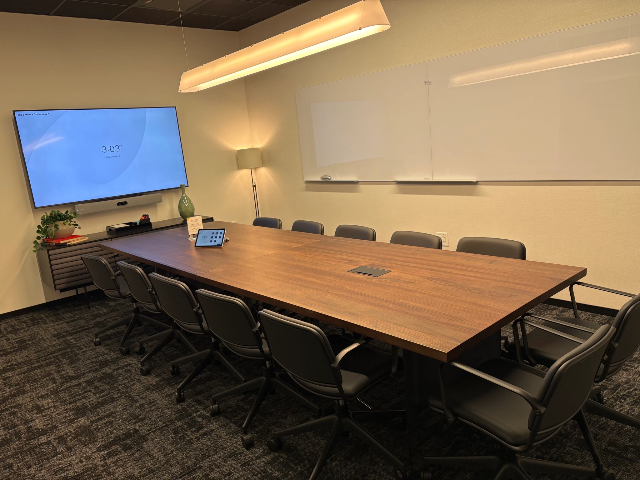 A modern coworking conference room in Charlotte featuring a long wooden table, ten chairs, a wall-mounted screen displaying the time, a whiteboard, and a side cabinet with plants and decor—ideal for your next workspace meeting.