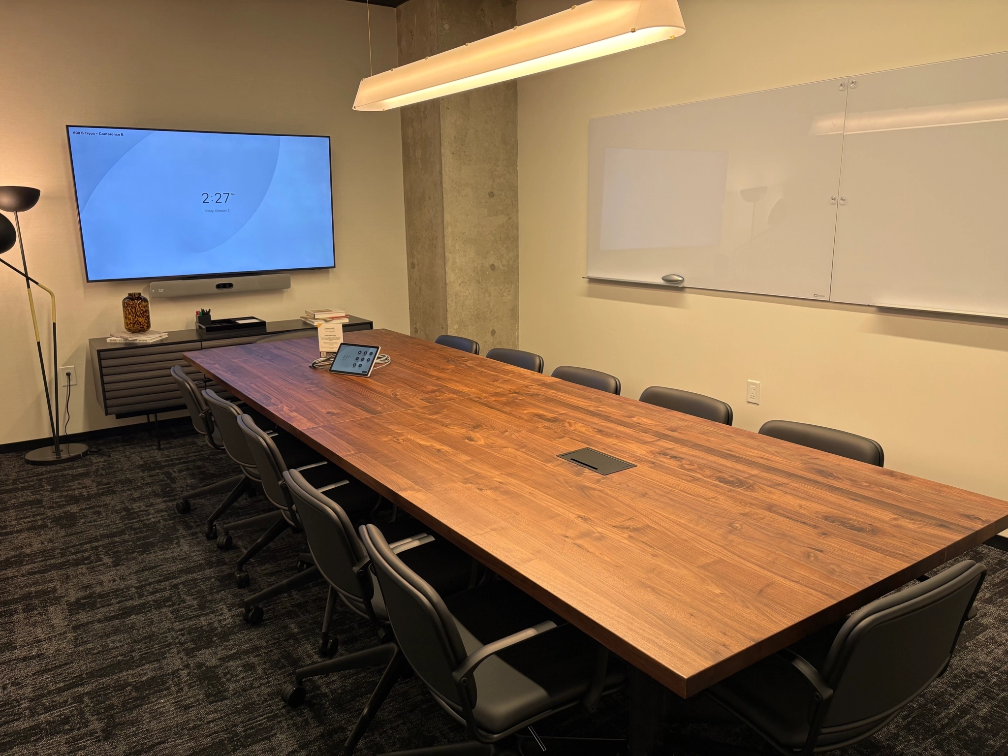 A modern meeting room with a long wooden table, ten chairs, a wall-mounted screen displaying the time, a whiteboard, and a ceiling light—ideal for coworking or office gatherings.