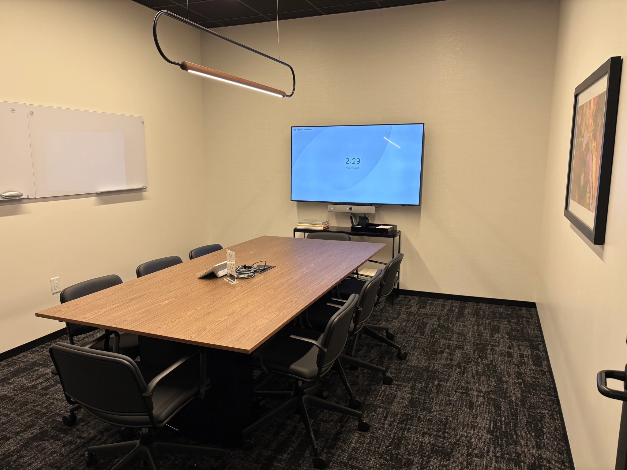 Small meeting room in a Charlotte office featuring a rectangular table, six chairs, a wall-mounted TV displaying 2:29, a whiteboard, and a framed picture on the wall.