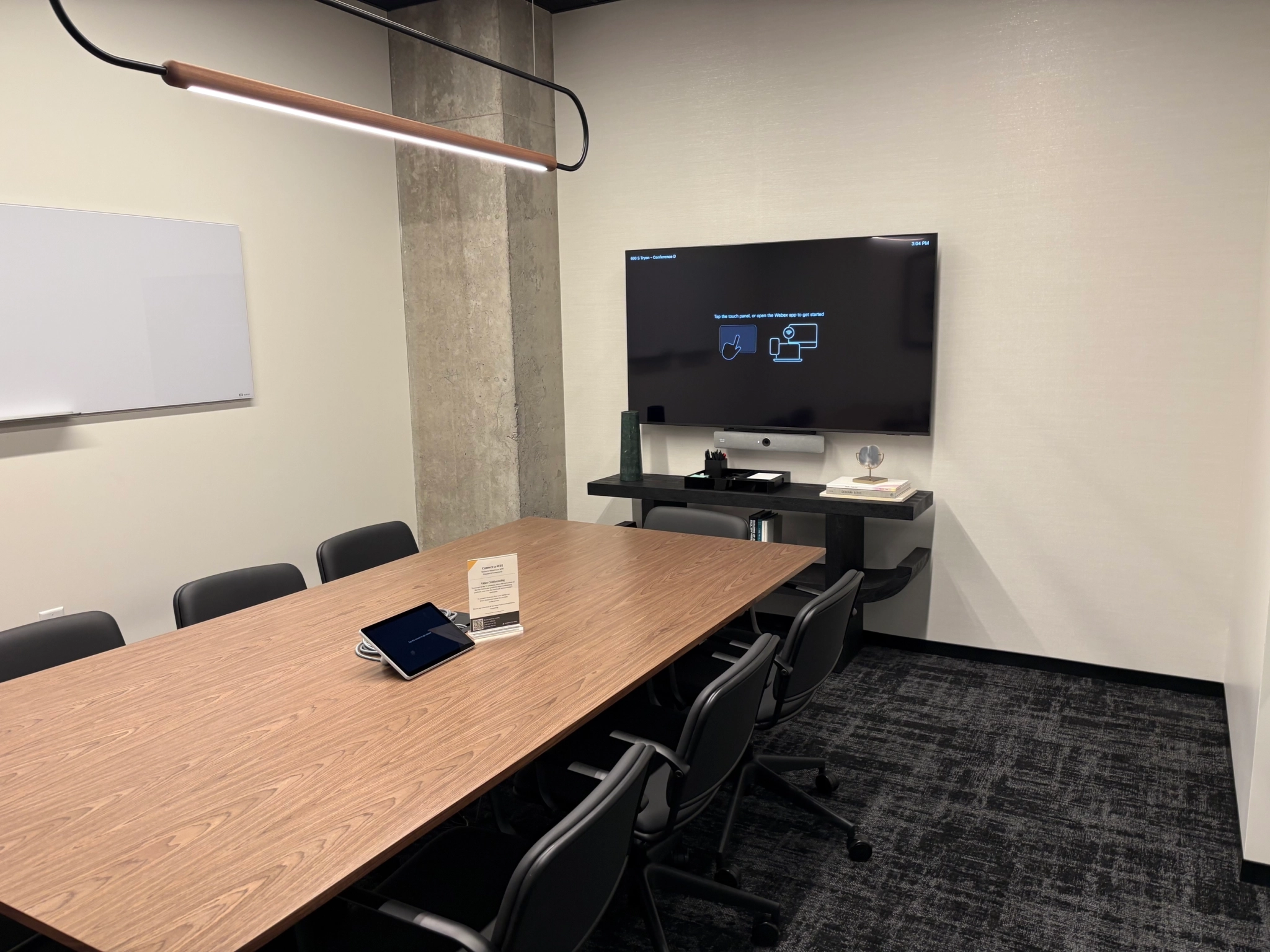 A modern meeting room in Charlotte features a long wooden table, several chairs, a wall-mounted monitor, a whiteboard, and an electronic device—ideal for coworking sessions and productive gatherings.