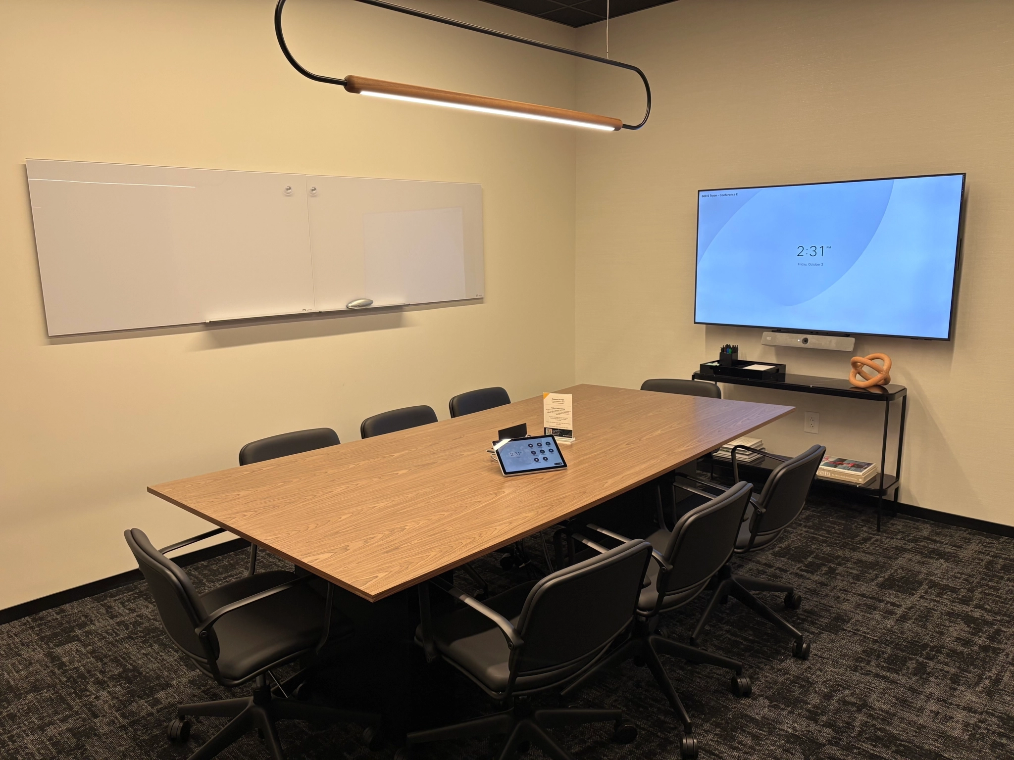 A modern meeting room with a wooden table, eight chairs, wall-mounted whiteboard, large screen display showing the time, and a touchscreen control device creates an efficient workspace for collaboration.
