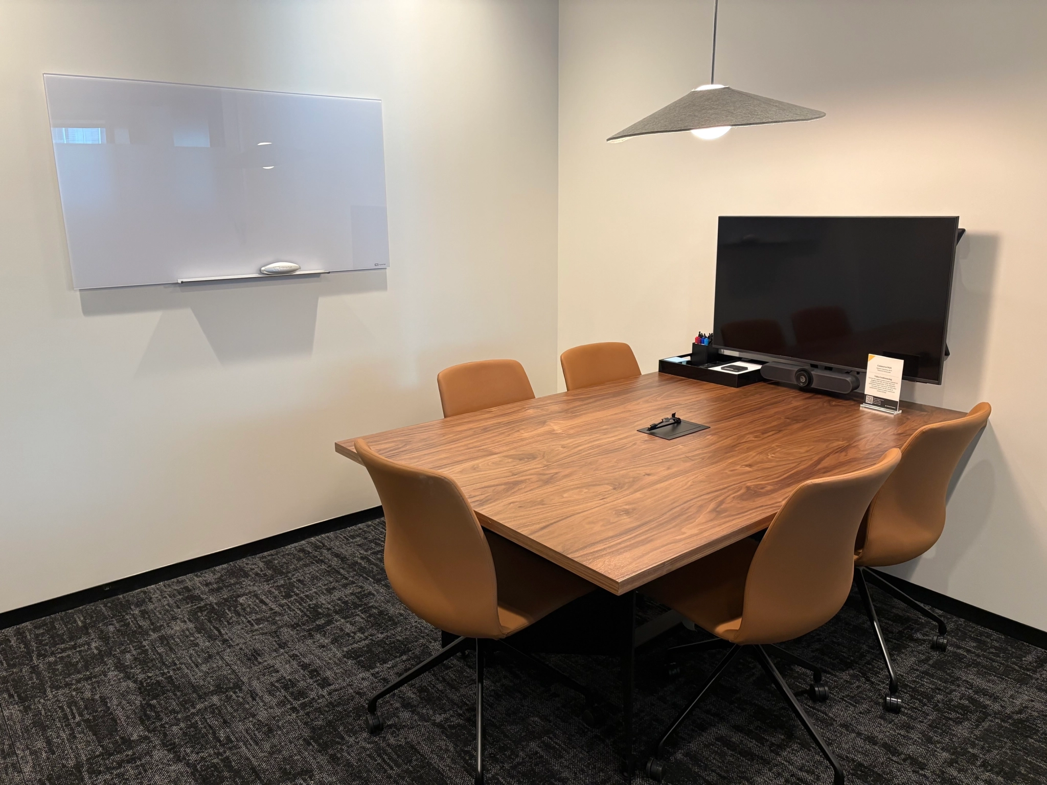 A small Charlotte meeting room with a wooden table, five tan chairs, a wall-mounted TV, a whiteboard, and a hanging light fixture—perfect for an intimate workspace.