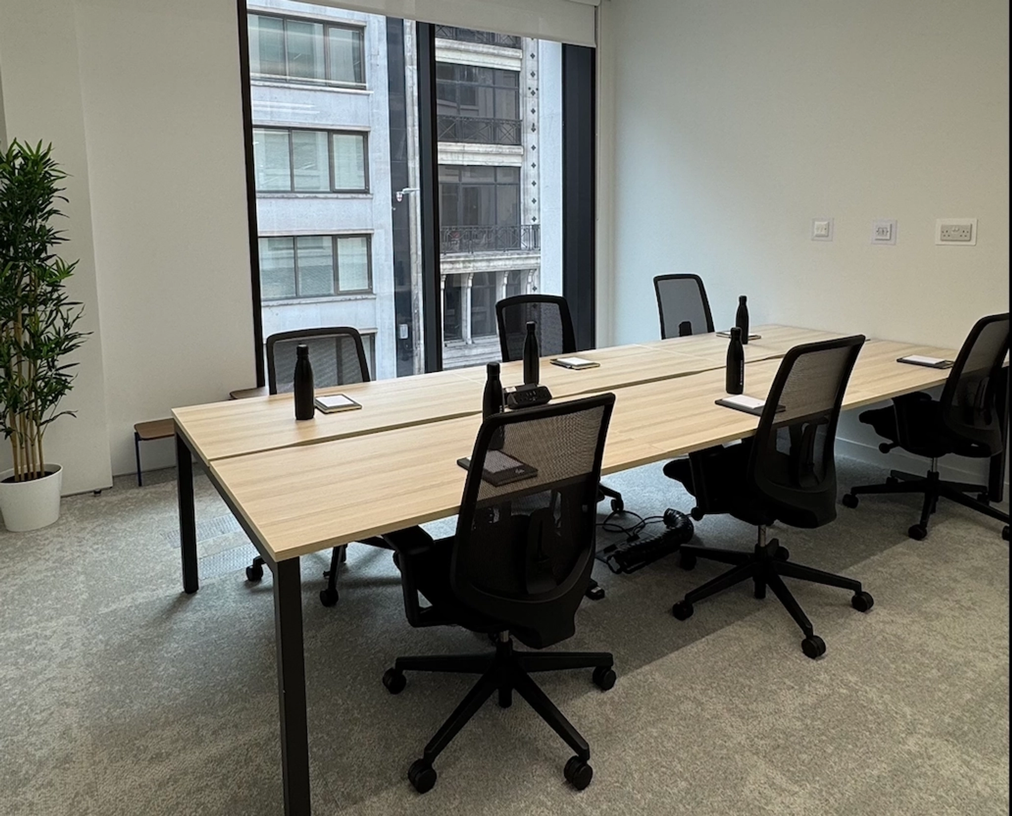A modern coworking meeting room with a light wood table, black chairs, water bottles, and notepads, set against large windows overlooking London city buildings.