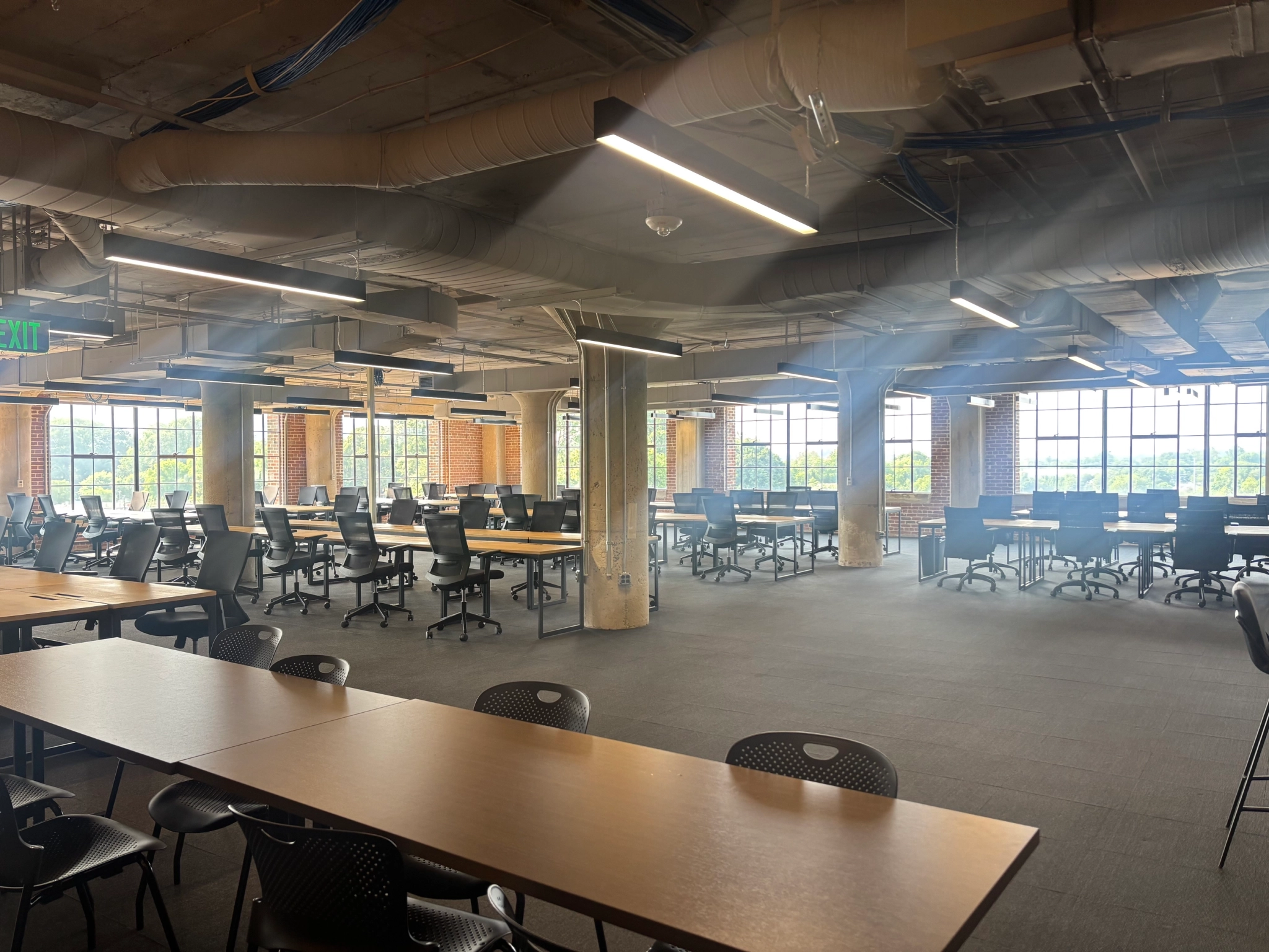 Spacious, modern workspace featuring rows of empty desks and chairs, exposed ceilings, brick walls, and sunlight pouring through large windows.