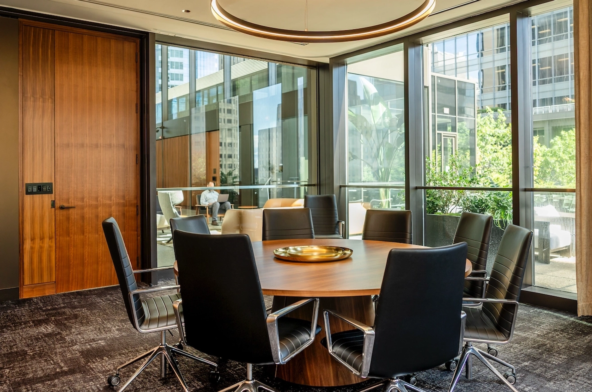 Modern coworking conference room with a round wooden table, eight black chairs, large windows, cityscape views, and abundant natural light.