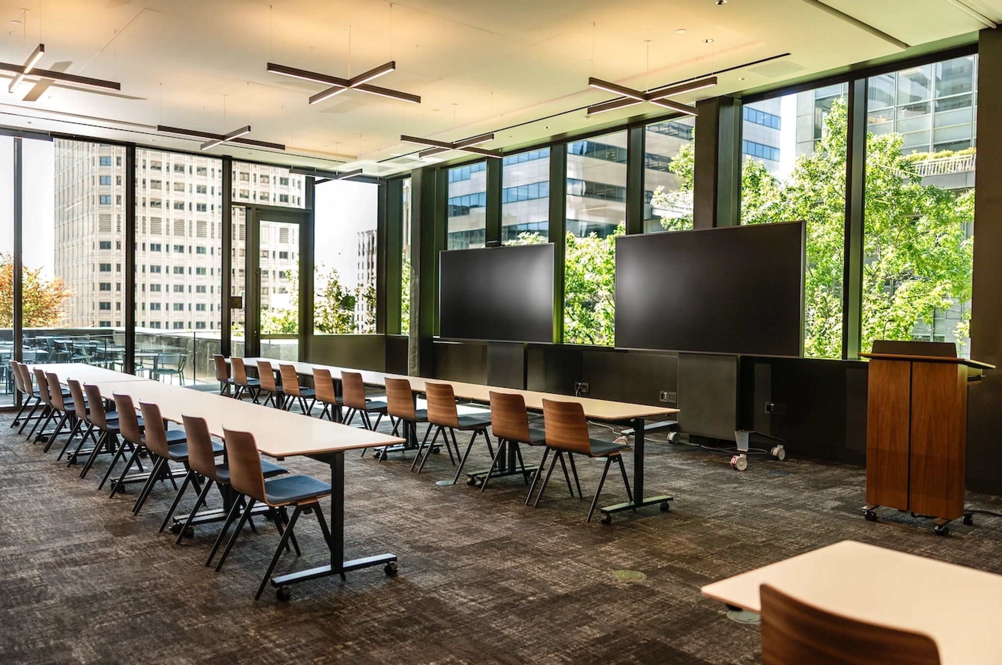 A modern coworking conference room in Seattle with rows of tables and chairs, two large screens at the front, floor-to-ceiling windows, and an empty podium. Perfect for your next workspace meeting or event.