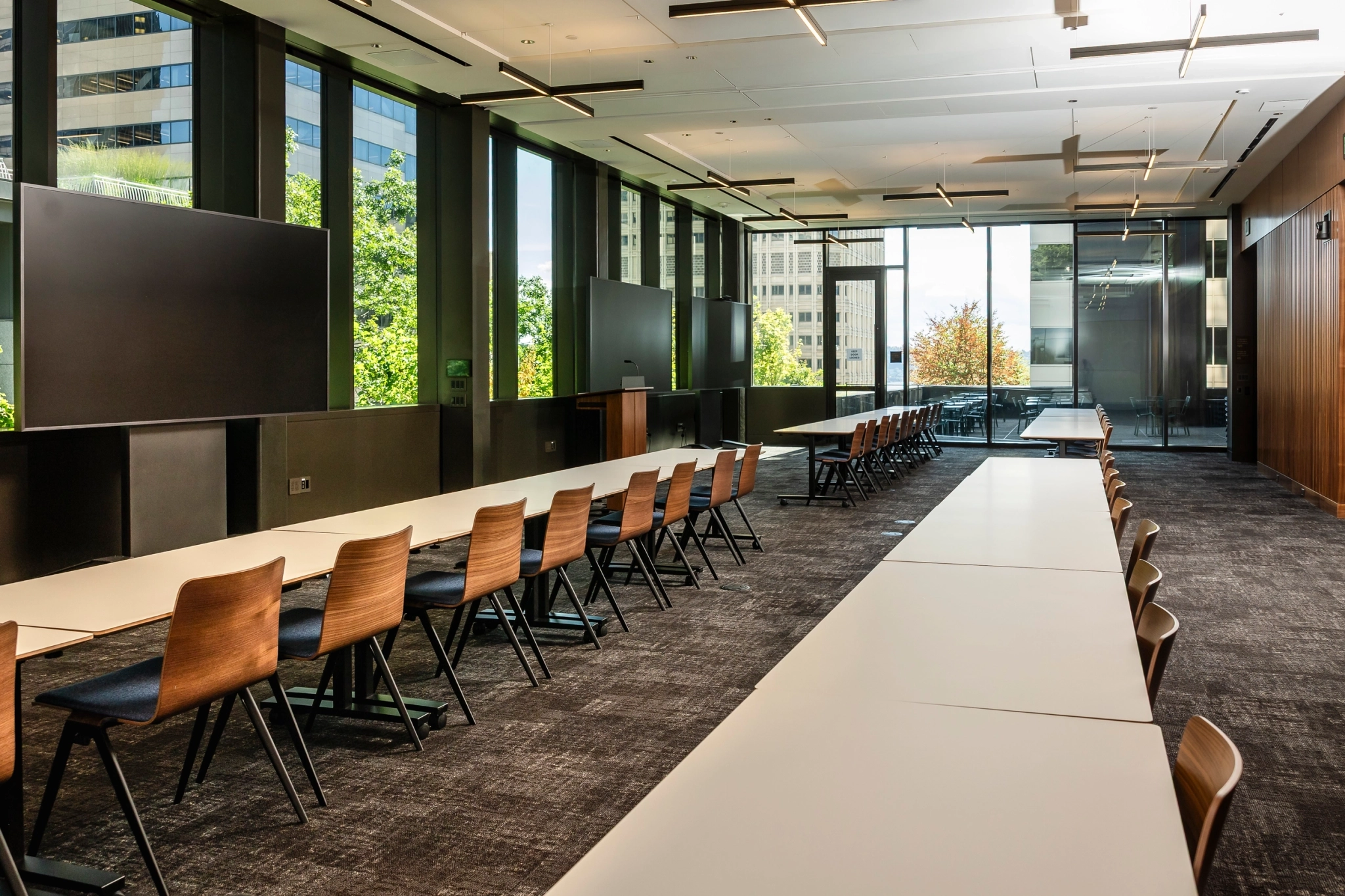Modern conference room with long tables, wooden chairs, large windows, and audio-visual screens; city buildings and greenery frame this inviting Seattle workspace.
