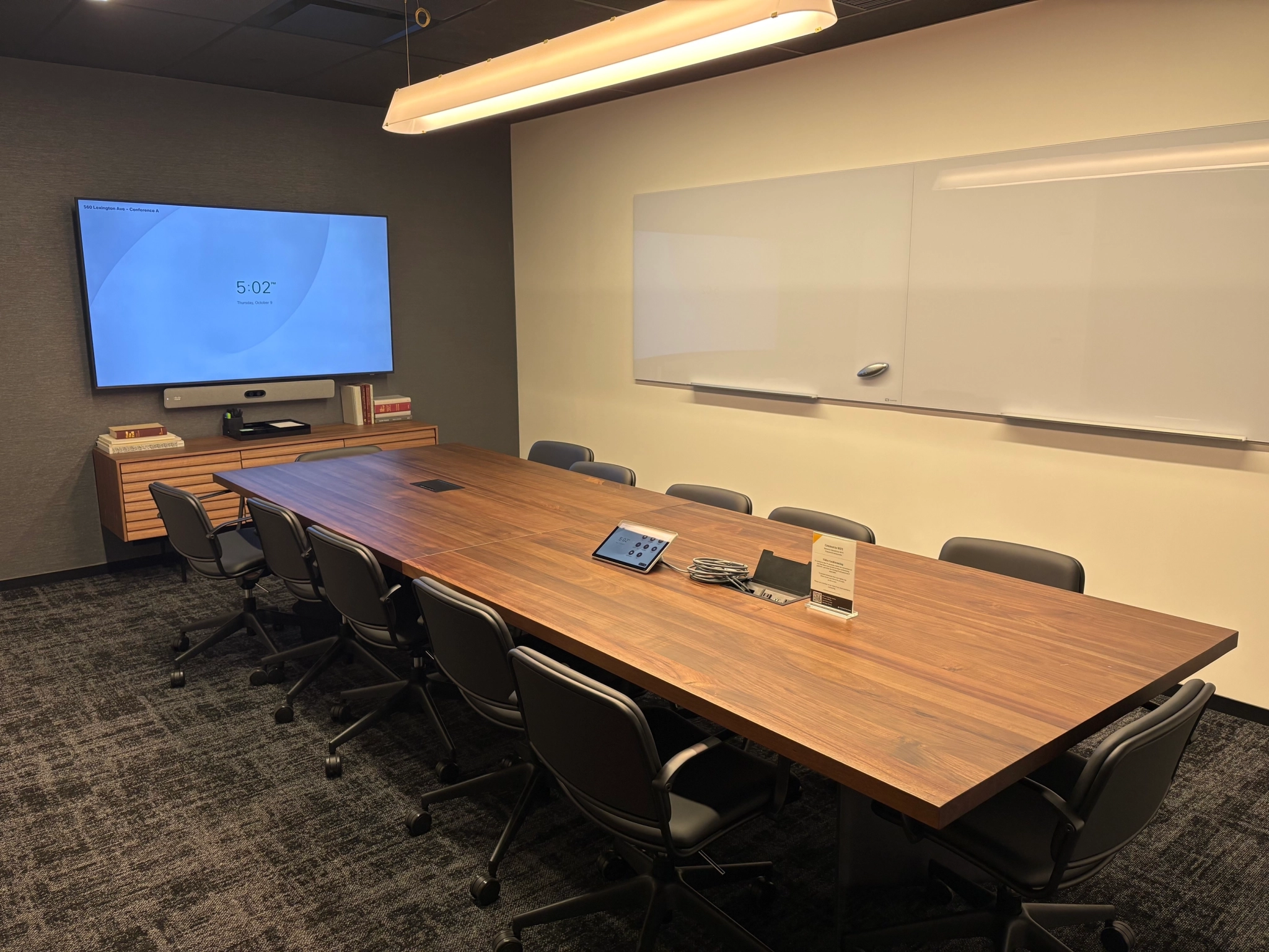 A modern meeting room in New York featuring a long wooden table, ten chairs, a wall-mounted screen, a whiteboard, and a small cabinet with books—perfect for collaborative office sessions.