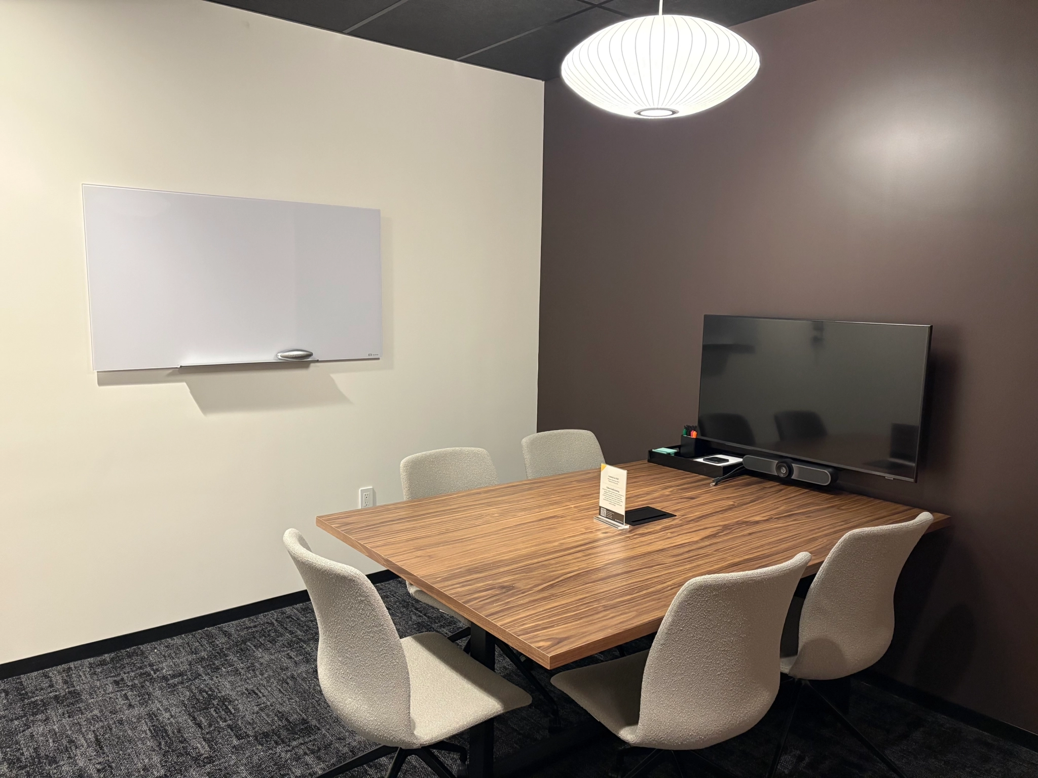 Small meeting room with a wooden table, six chairs, a wall-mounted TV, a whiteboard, and a hanging light fixture—ideal for office or coworking sessions.
