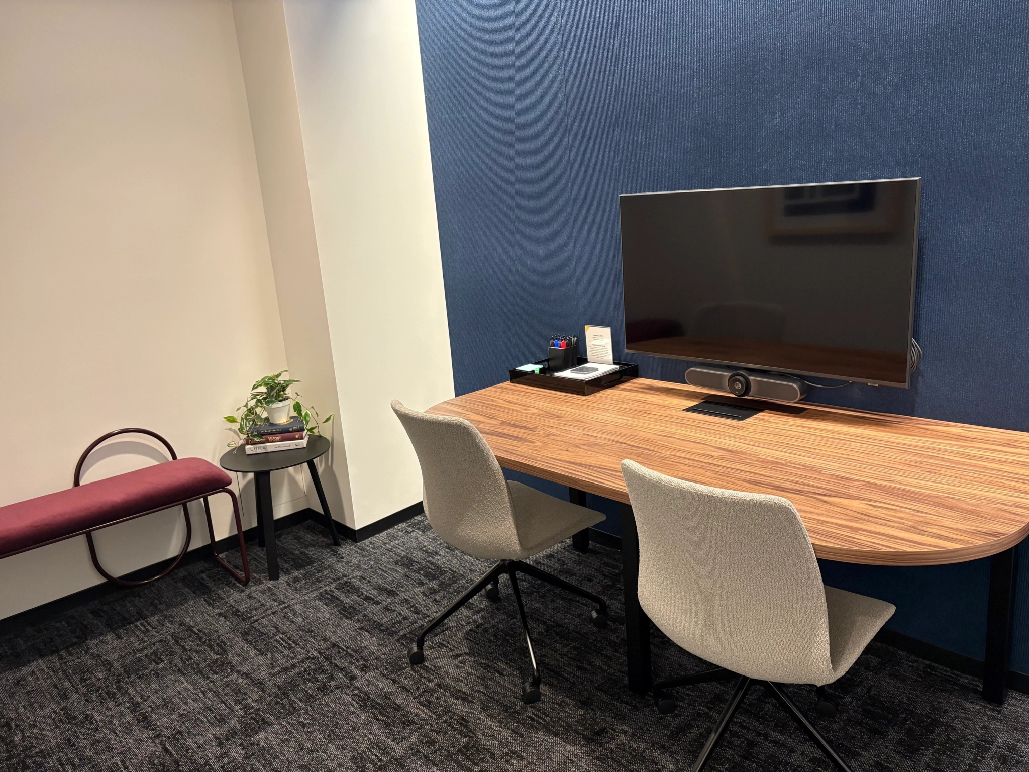 A small coworking office with a wooden desk, two chairs, a large monitor, a side table with books and a plant, and a maroon bench against a blue accent wall—perfect for a productive day in New York.