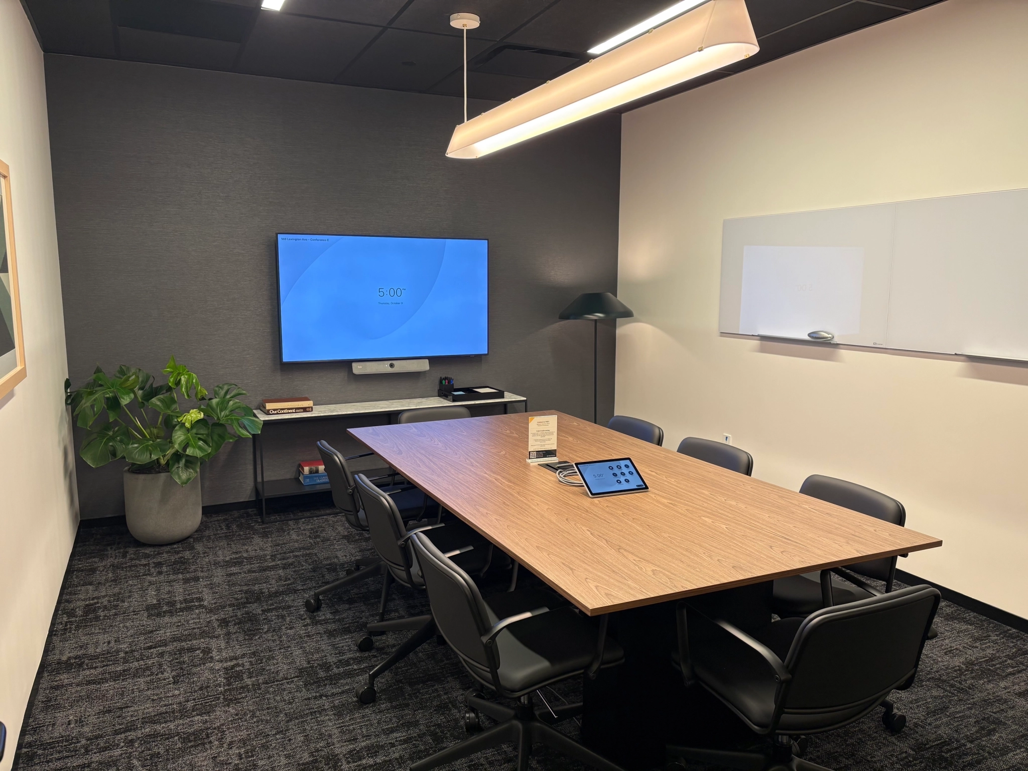 A modern meeting room with a wooden table, eight chairs, a large screen on the wall, whiteboards, a potted plant, and a ceiling light—perfect for any office workspace.