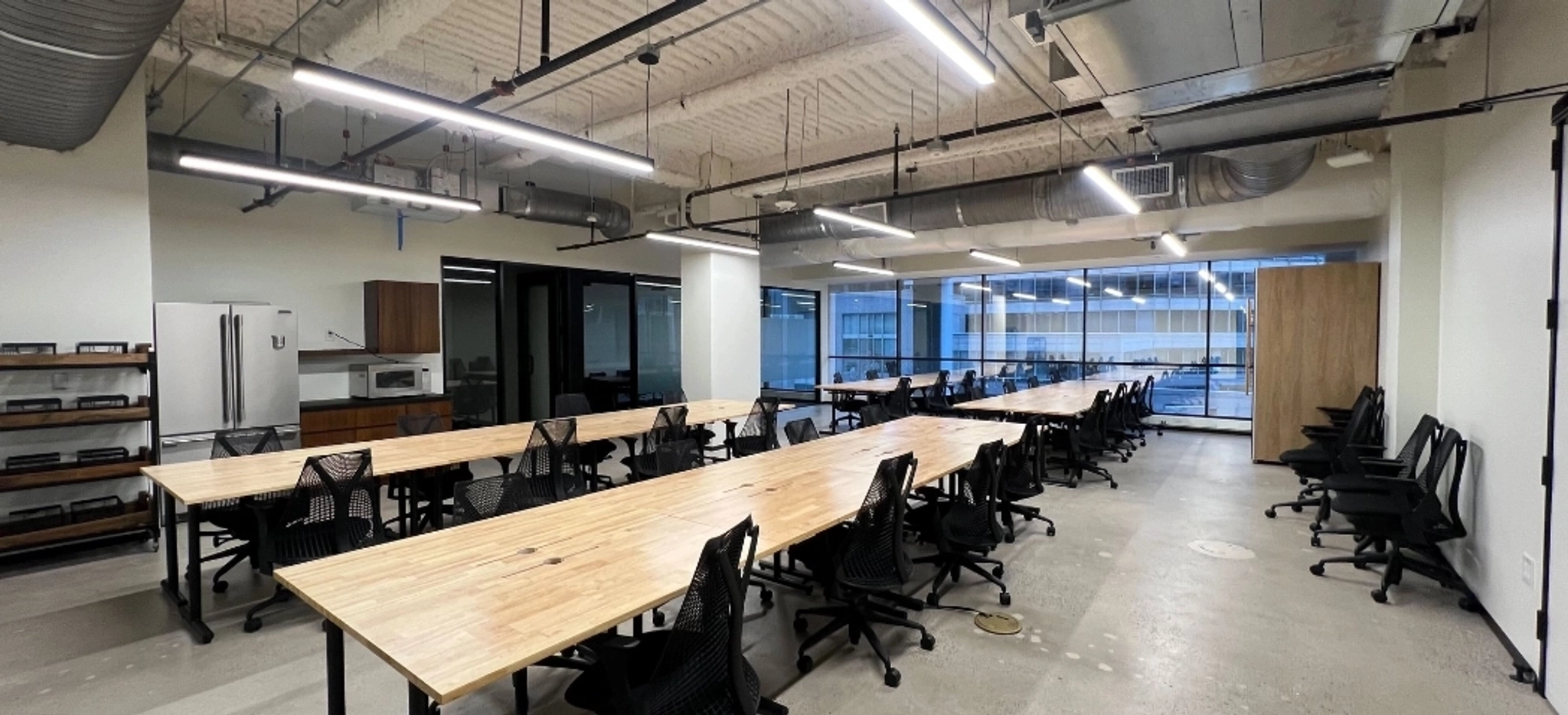 Modern open-plan workspace with long wooden desks, black mesh chairs, overhead lighting, exposed ceiling, kitchen appliances, and large windows along the back wall. Perfect for coworking and collaborative projects.