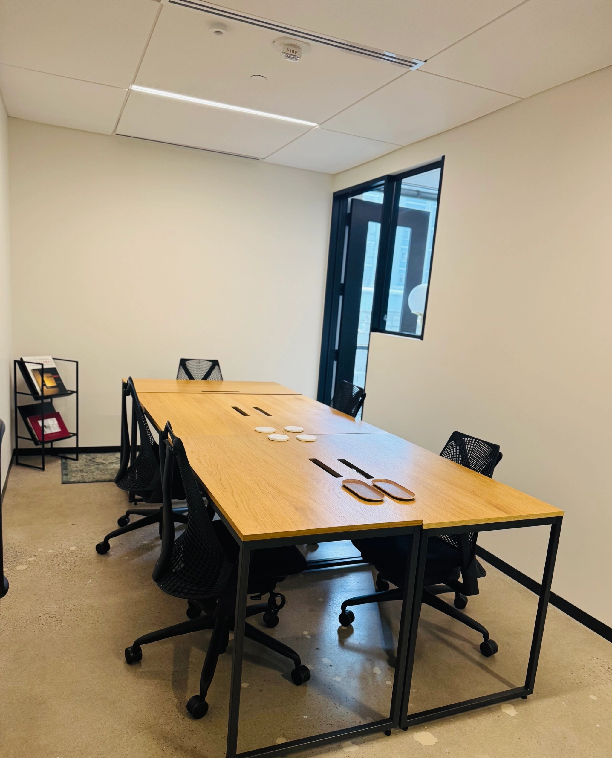 A modern office meeting room with a wooden table, six black chairs, coasters, scissors, a stapler, and a bookshelf with folders near the wall.